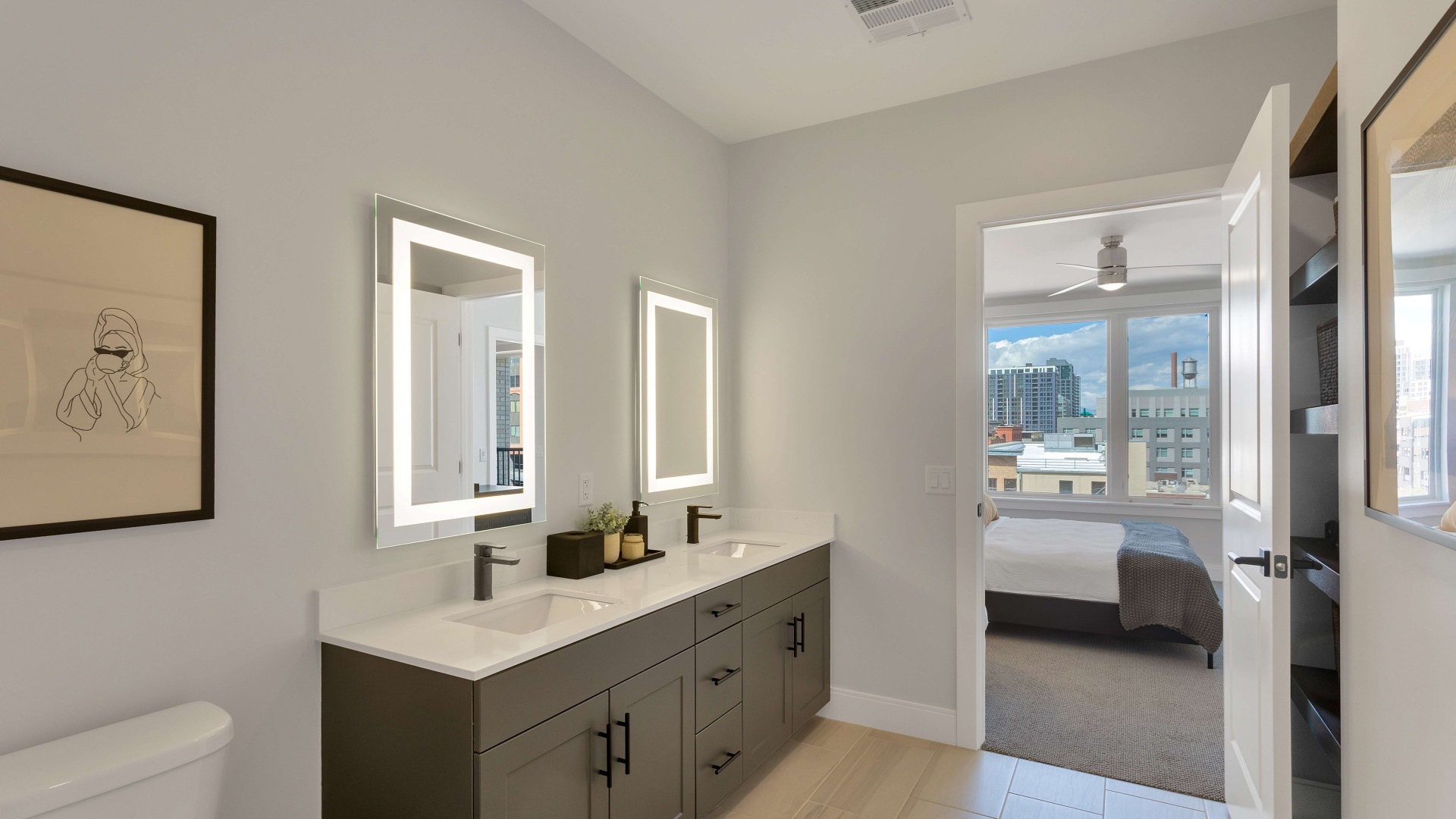 Modern bathroom with dual sinks and illuminated mirrors, flowing into a bright bedroom with large windows offering city views.	 Modern bathroom with dual sinks and illuminated mirrors, flowing into a bright bedroom with large windows offering city views.
